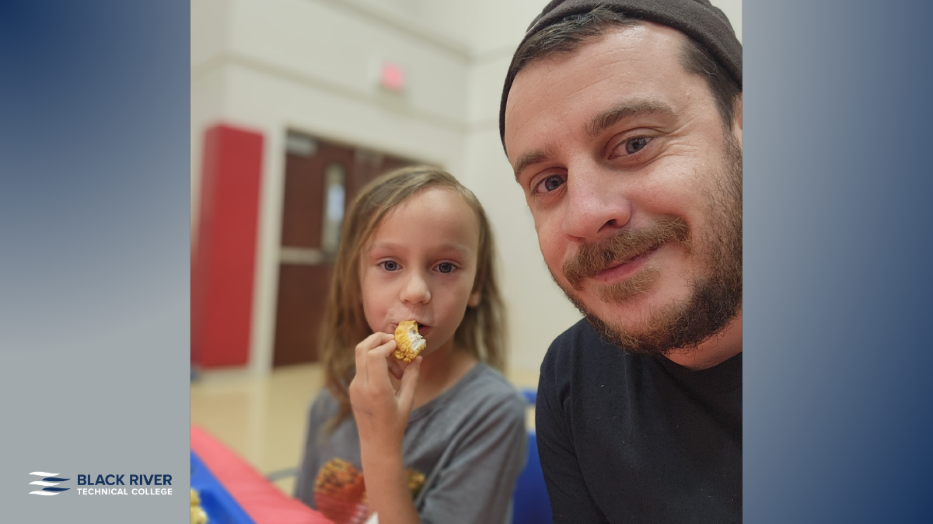 A man in a black beanie and shirt takes a selfie with a young girl beside him, both holding food, in an indoor gymnasium setting with Black River Technical College branding.